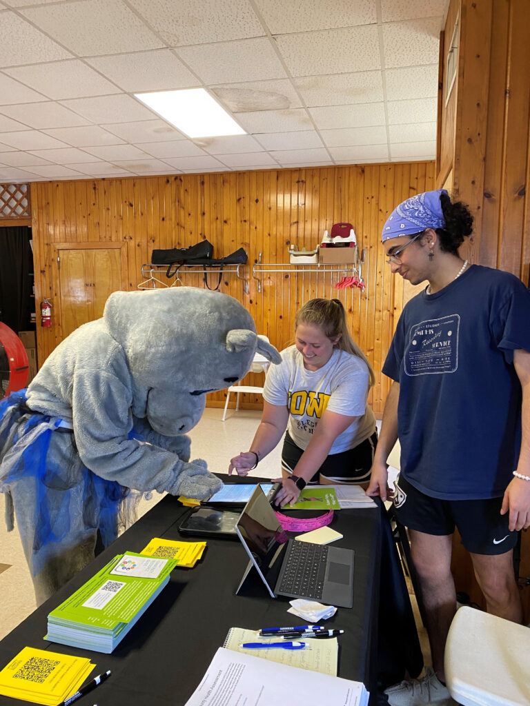 Two students with the CPH Strike Force help a hippo mascot at a table.