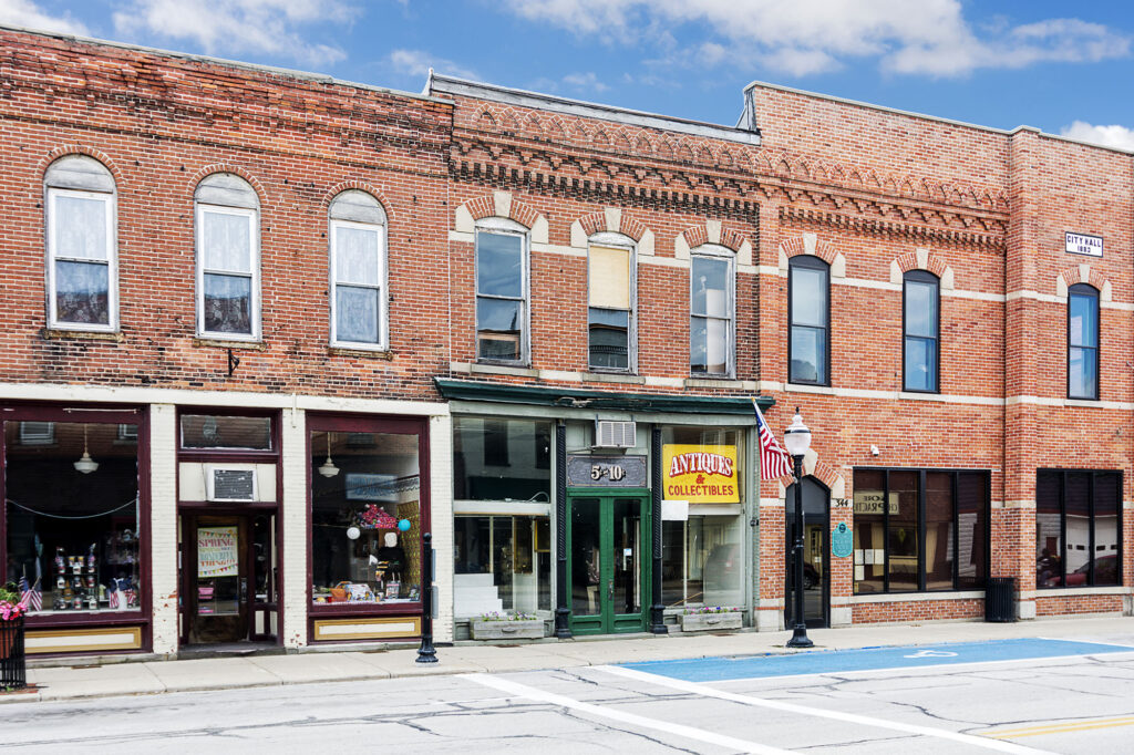 A photo of a typical small town main street in the United States of America. Features old brick buildings with specialty shops and restaurants including an old five and dime store. Decorated with spring flowers and American flags.