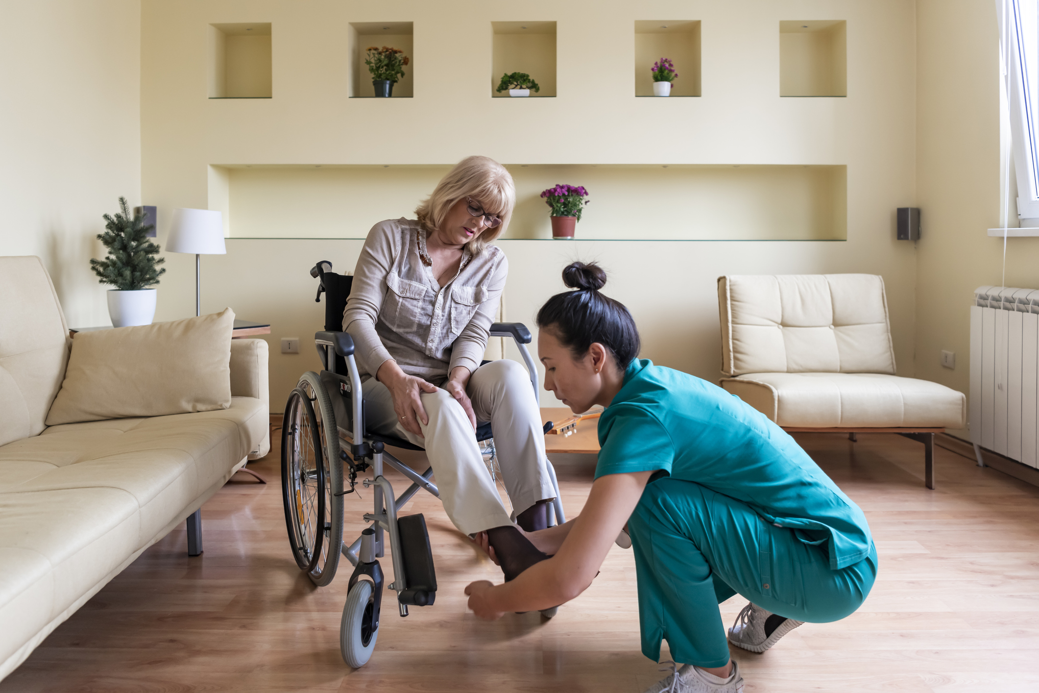 Sick Old Woman in a Wheelchair is Receiving a Help From a Young Nurse in Home Visit.